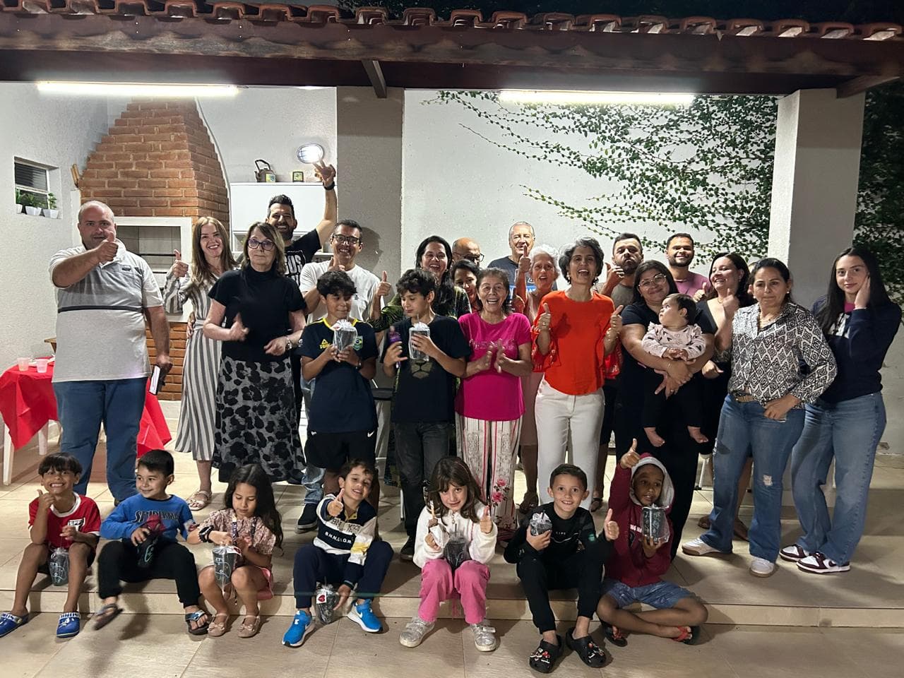Célula reunida na varanda de uma casa à noite, sob um varal de luzinhas, com as crianças sentadas na frente segurando copos