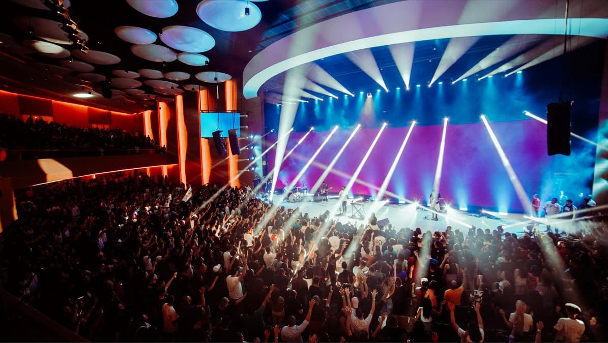 Auditório lotado num culto da Videira, com a plateia de mãos levantadas diante do palco iluminado
