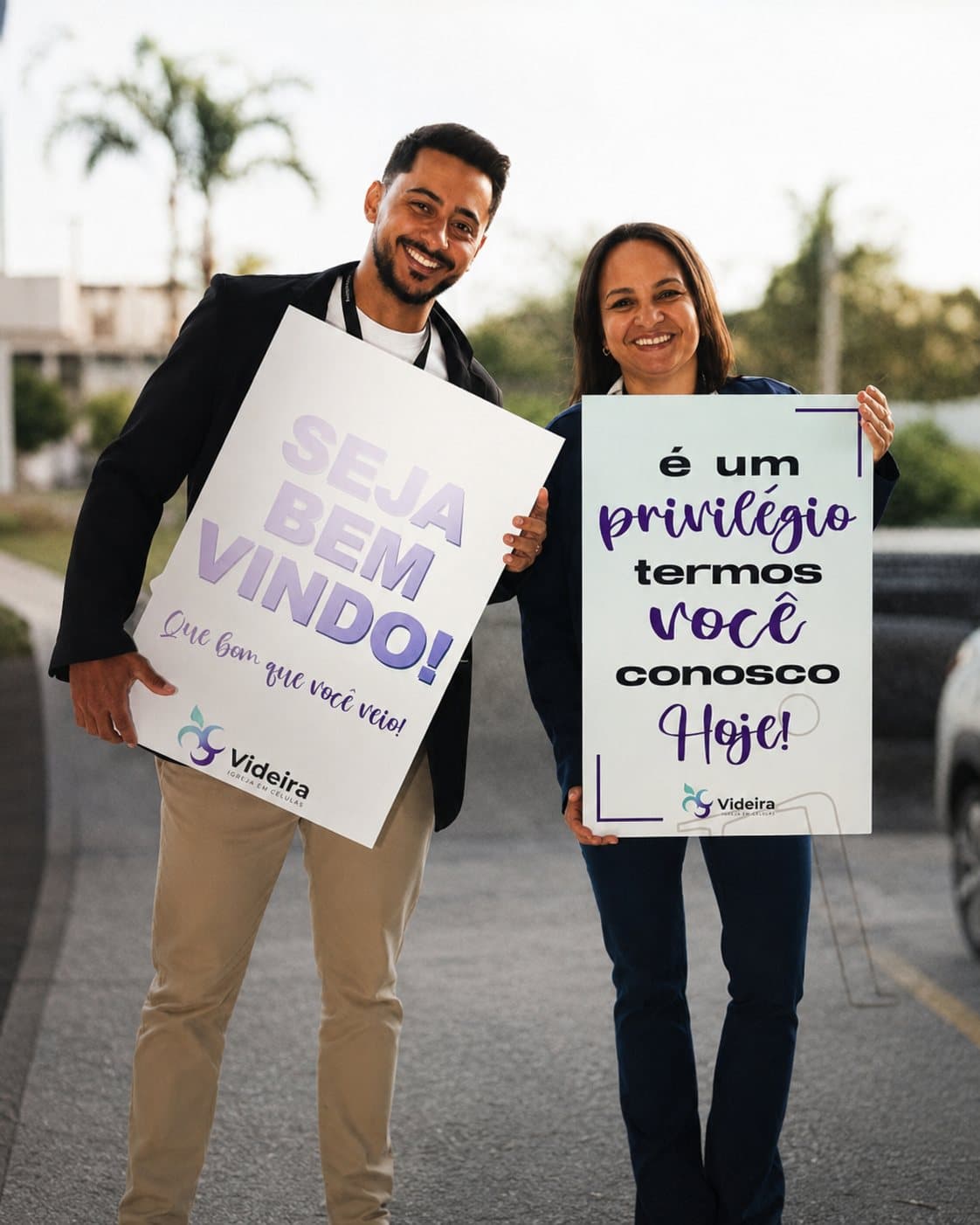 Dois voluntários da Videira recebendo visitantes com placas escritas 'Seja bem-vindo' e 'É um privilégio termos você conosco hoje'