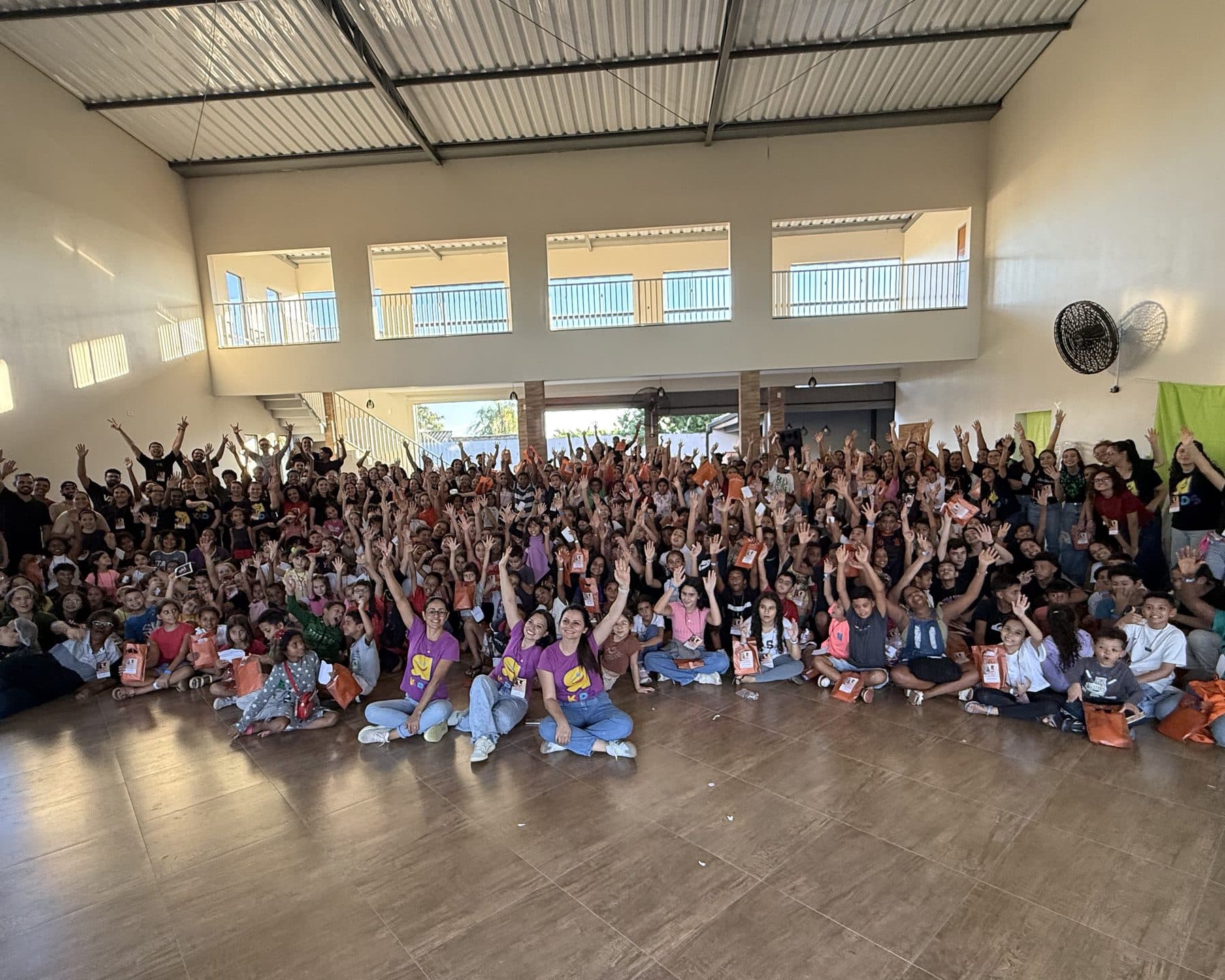 Salão cheio de crianças com os braços erguidos num evento do Radicais Kids