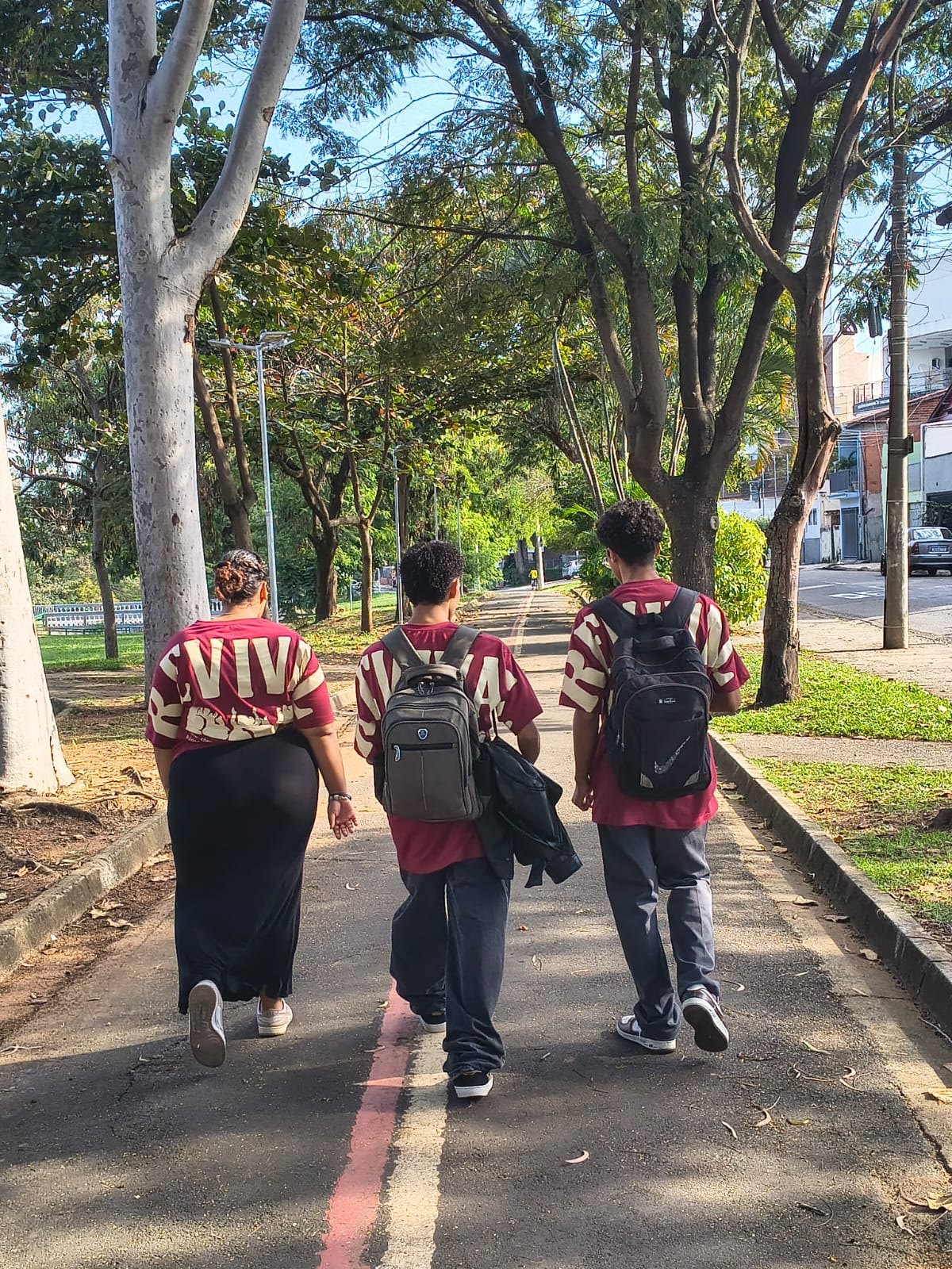 Três jovens de mochila caminhando de costas por uma ciclovia arborizada