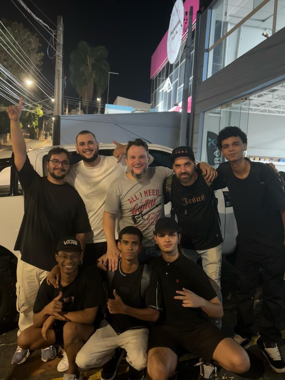 Grupo de rapazes posando na calçada à noite em frente a uma loja iluminada, um deles com o braço erguido